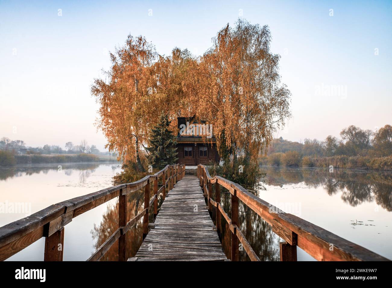 Paesaggio mattutino di una piccola casa su un'isola artificiale con un lungo ponte di legno. Intorno alla casa c'è un piccolo lago con nebbia leggera. Paesaggio autunnale Foto Stock