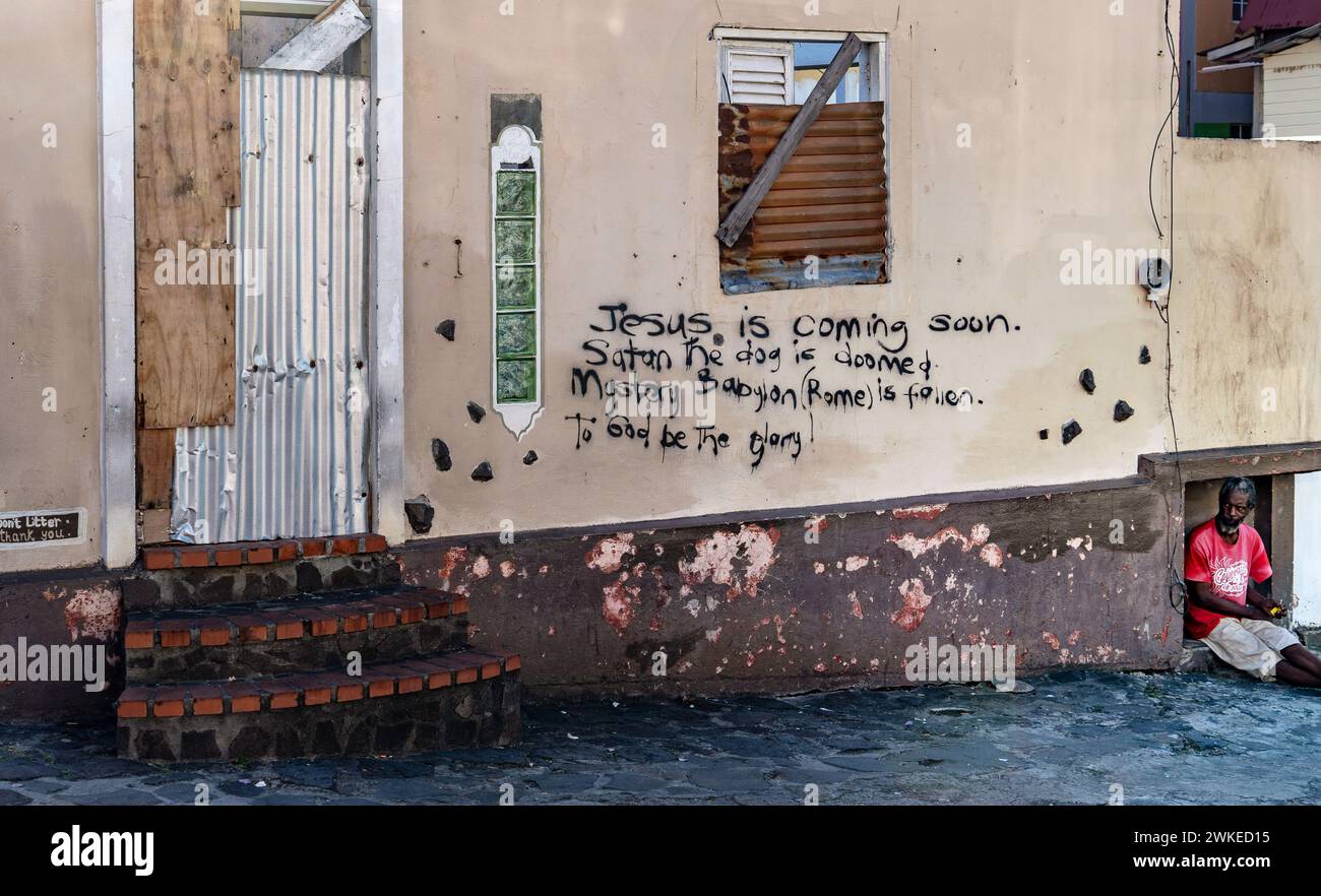 Un mendicante seduto davanti a un muro con una scritta su Gesù che verrà presto e Satana che sarà condannato Foto Stock