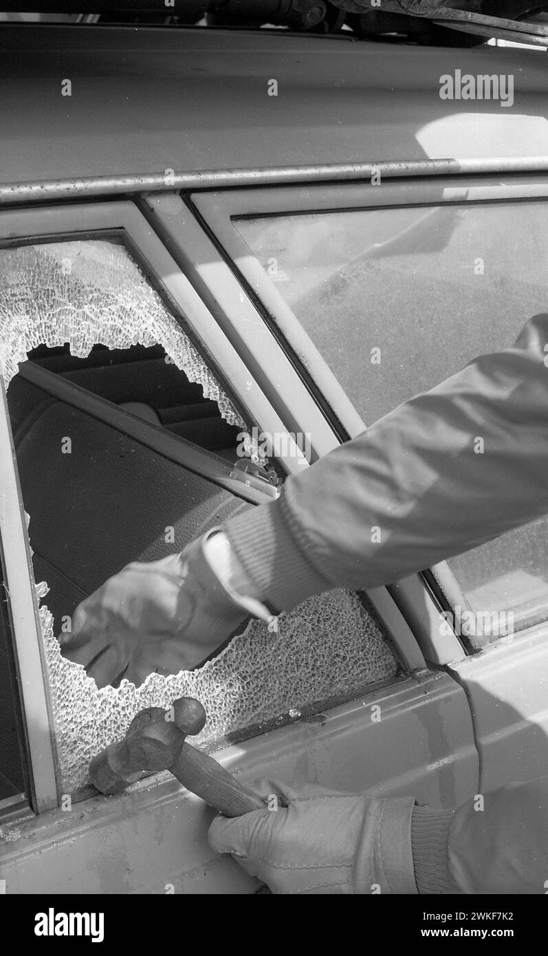 Anni '1970, storico, furto d'auto, una mano di un guanto che butta fuori il vetro frantumato del finestrino laterale di un'auto, un martello tenuto in un'altra mano con i guanti, Inghilterra, Regno Unito. Foto Stock