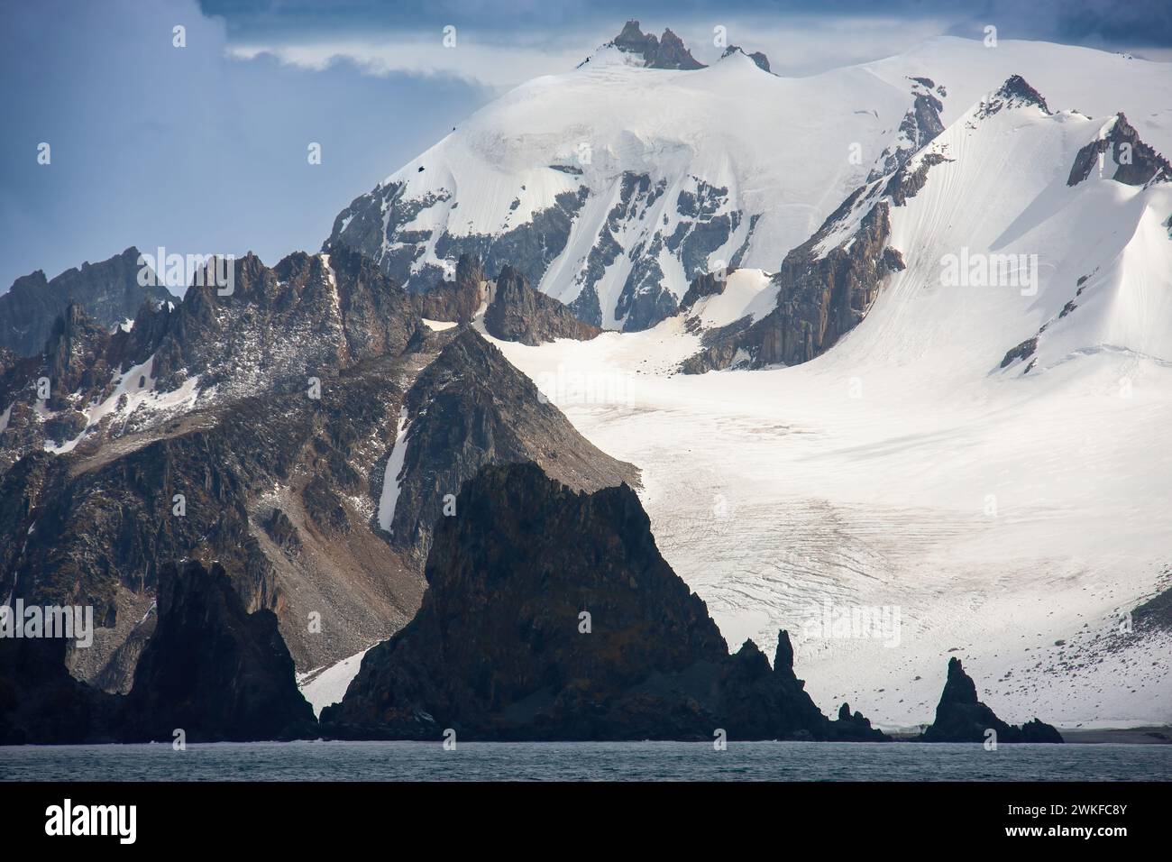 Un ampio affioramento di formazioni basaltiche che si innalzano fuori da una baia sullo sfondo di alte cime di montagna e di un ghiacciaio. Penisola Antartica, Antartide Foto Stock