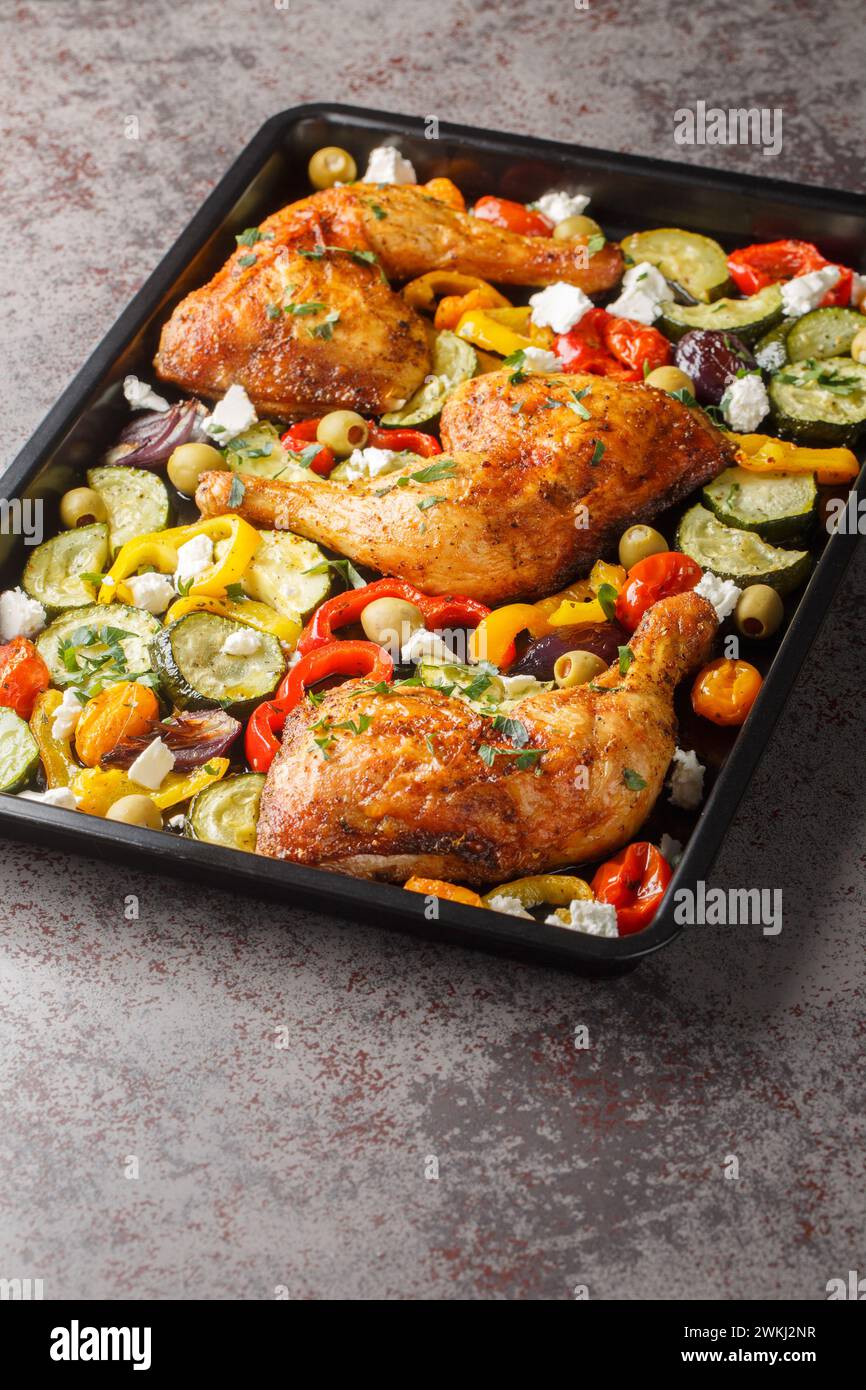 Cosce di pollo al forno con zucchine, pomodori, peperoni, olive e formaggio feta in primo piano su una teglia da forno sul tavolo. Verticale Foto Stock