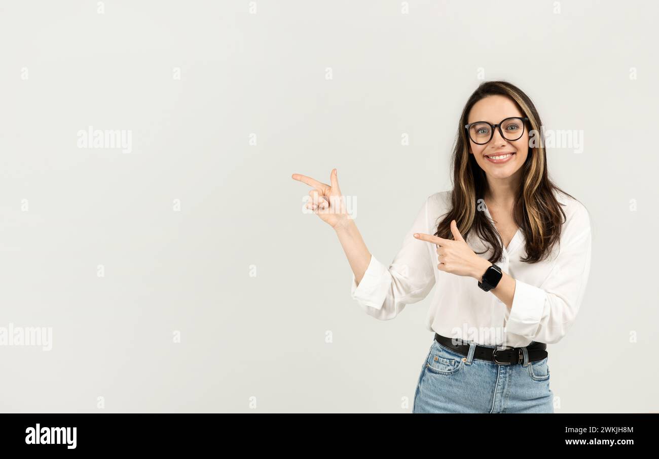 Donna felice con occhiali che puntano le dita verso un lato, indossando una camicia bianca e jeans Foto Stock
