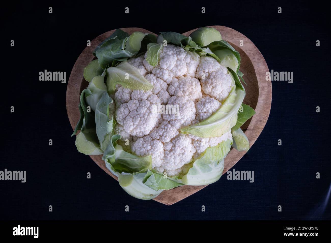 Cavolfiore organico fresco in un piatto di legno a forma di cuore su sfondo nero Foto Stock