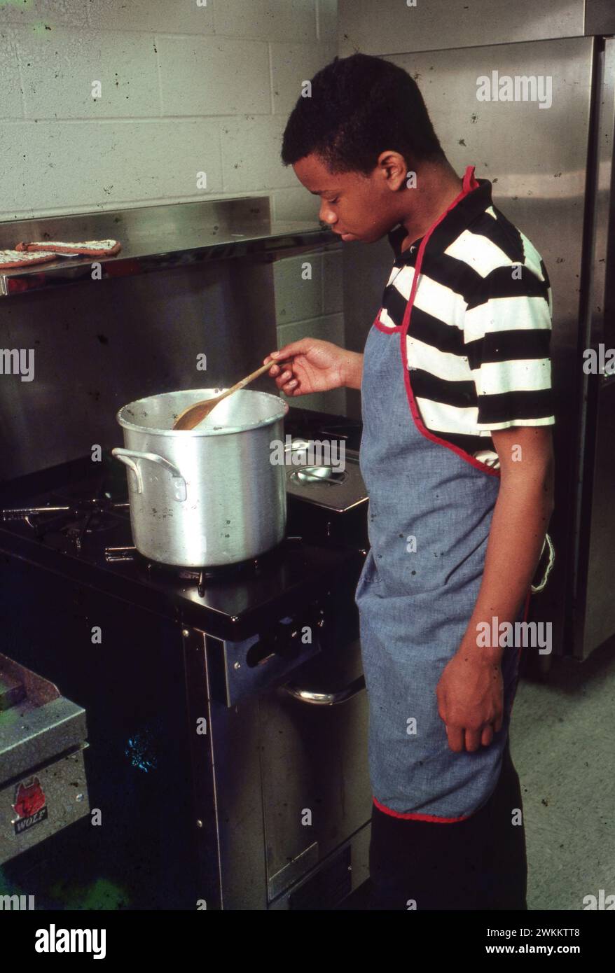 Un adolescente di colore che indossa un grembiule mescola il cibo sul piano cottura in classe di preparazione professionale. ©Bob Daemmrich Foto Stock