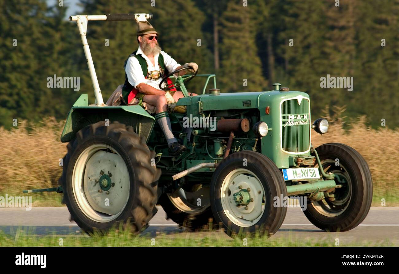 Contadino vestito tradizionalmente in lederhosen e cappello tirolese su un trattore, Dietramszell, Baviera, Germania Foto Stock