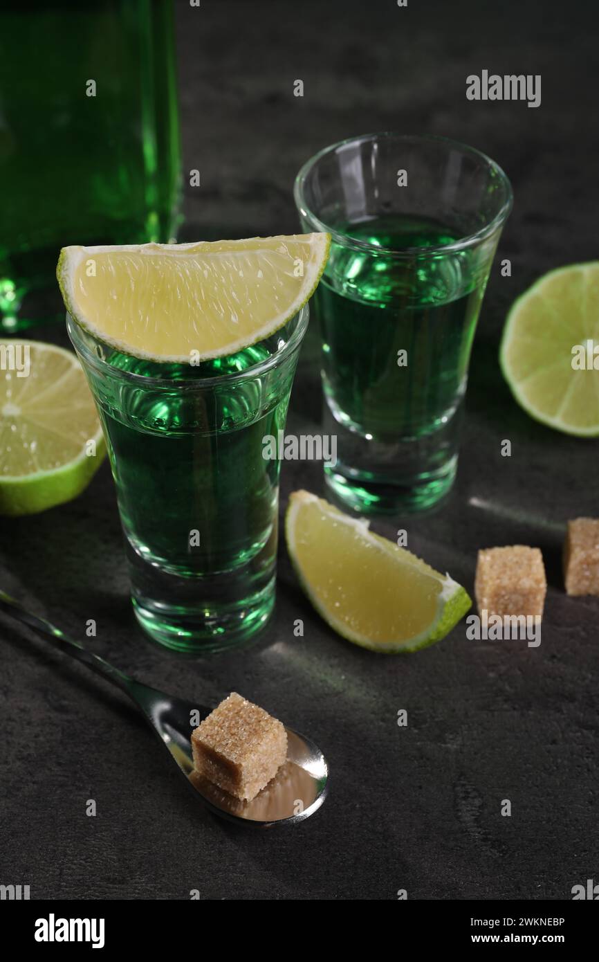 Absinthe in bicchieri da shot, cucchiaio, cubetti di zucchero di canna e lime su un tavolo testurizzato grigio, primo piano. Bevanda alcolica Foto Stock