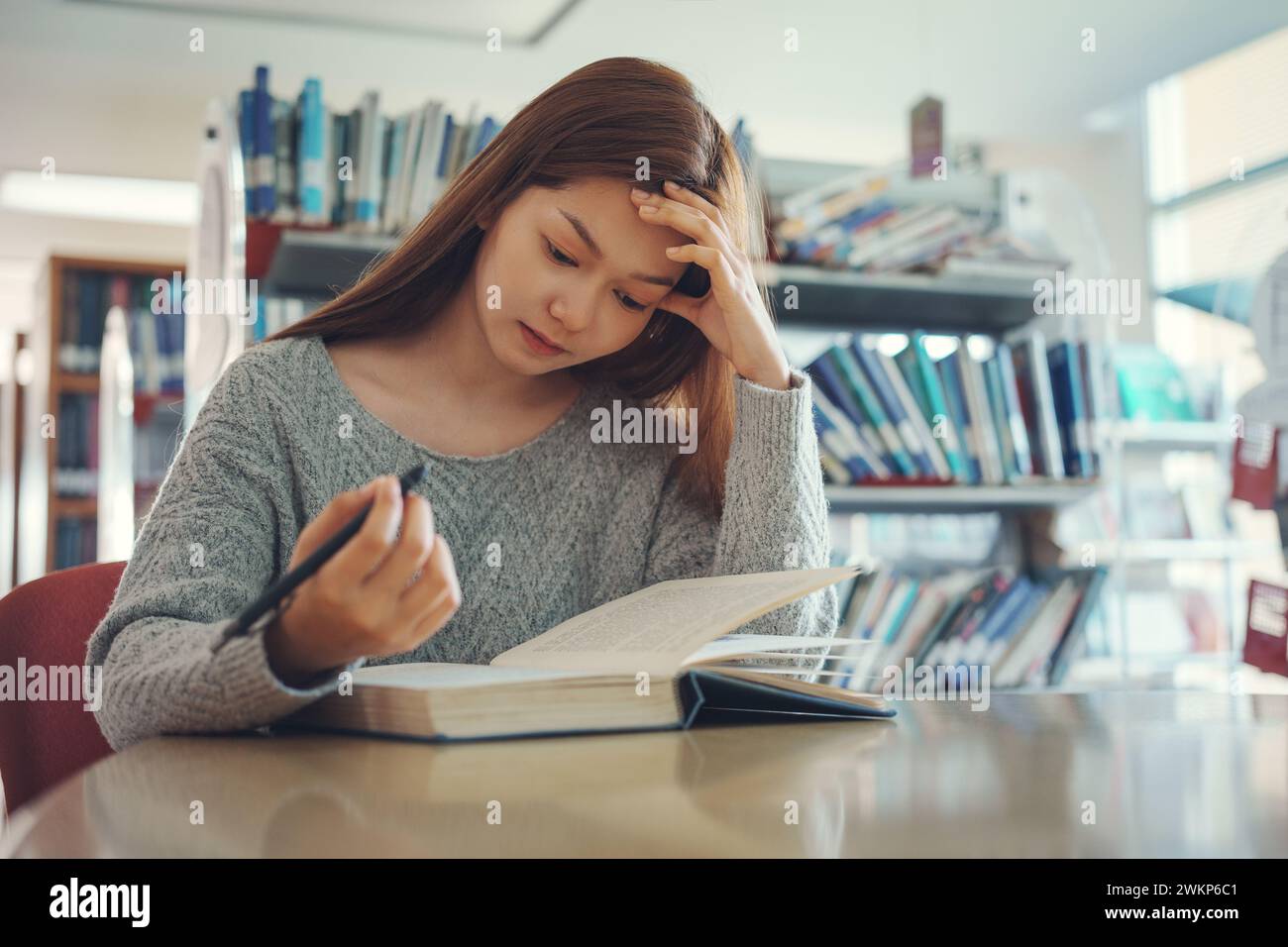 Una giovane studentessa stressata seduta a un tavolo di legno e a leggere un libro durante la preparazione agli esami in biblioteca. Foto Stock