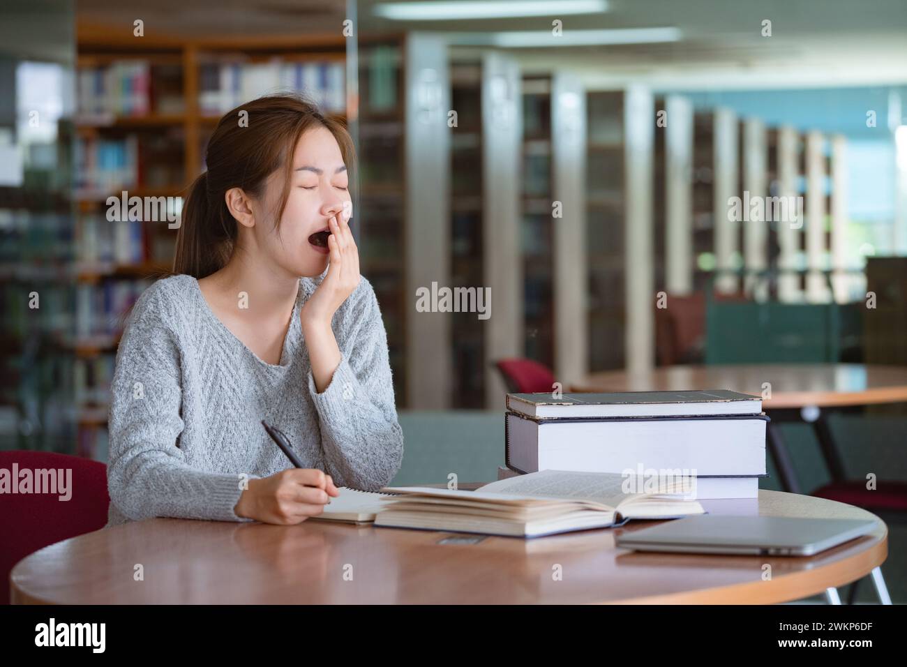 Giovane donna asiatica che sbadiglia mentre legge un libro in biblioteca. Foto Stock