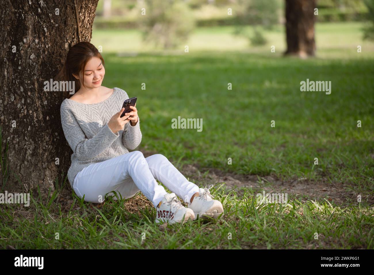 Giovane donna asiatica che usa lo smartphone mentre siede a terra nel parco cittadino. Foto Stock