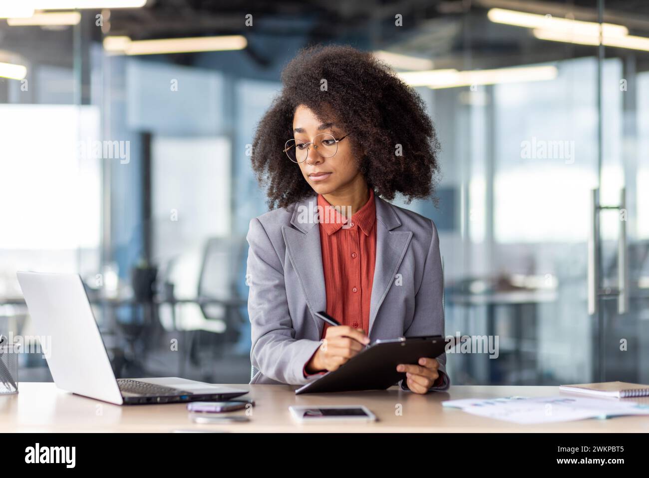 Donna concentrata con occhiali rotondi che guarda lo schermo del pc mentre compila il documento su una lavagna negli interni dell'ufficio. Imprenditore sicuro che copia le informazioni da e-mail a documenti cartacei. Foto Stock