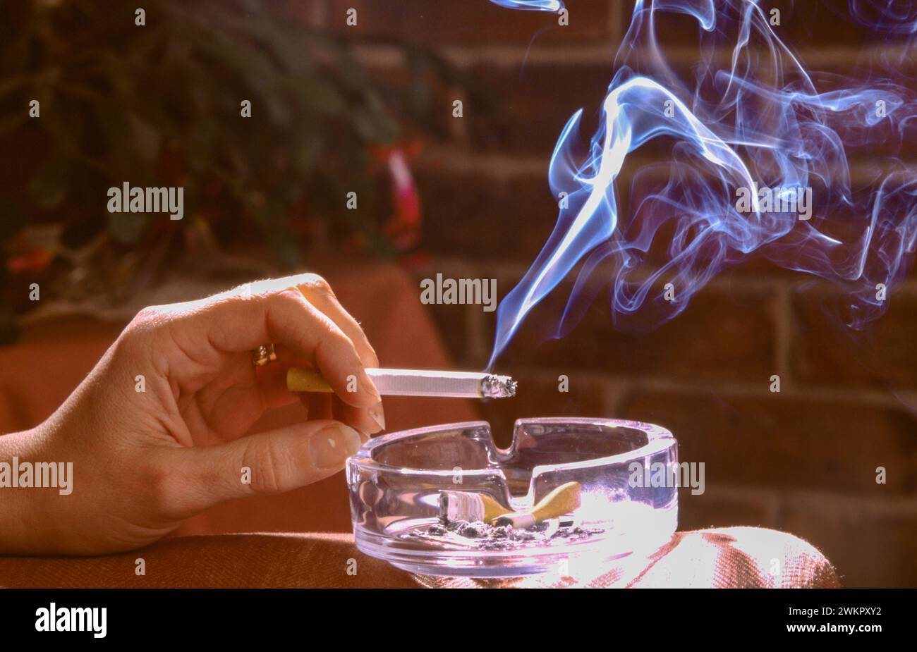 mano che tiene una sigaretta su un posacenere con il fumo che sale in una stanza Foto Stock