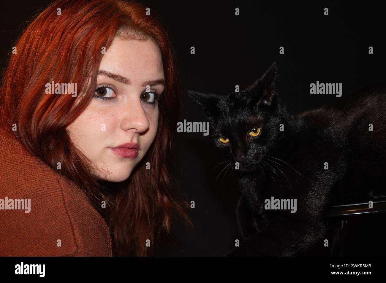 Una ragazza dai capelli rossi con il suo gatto nero e sfondo nero, entrambi guardano avanti Foto Stock