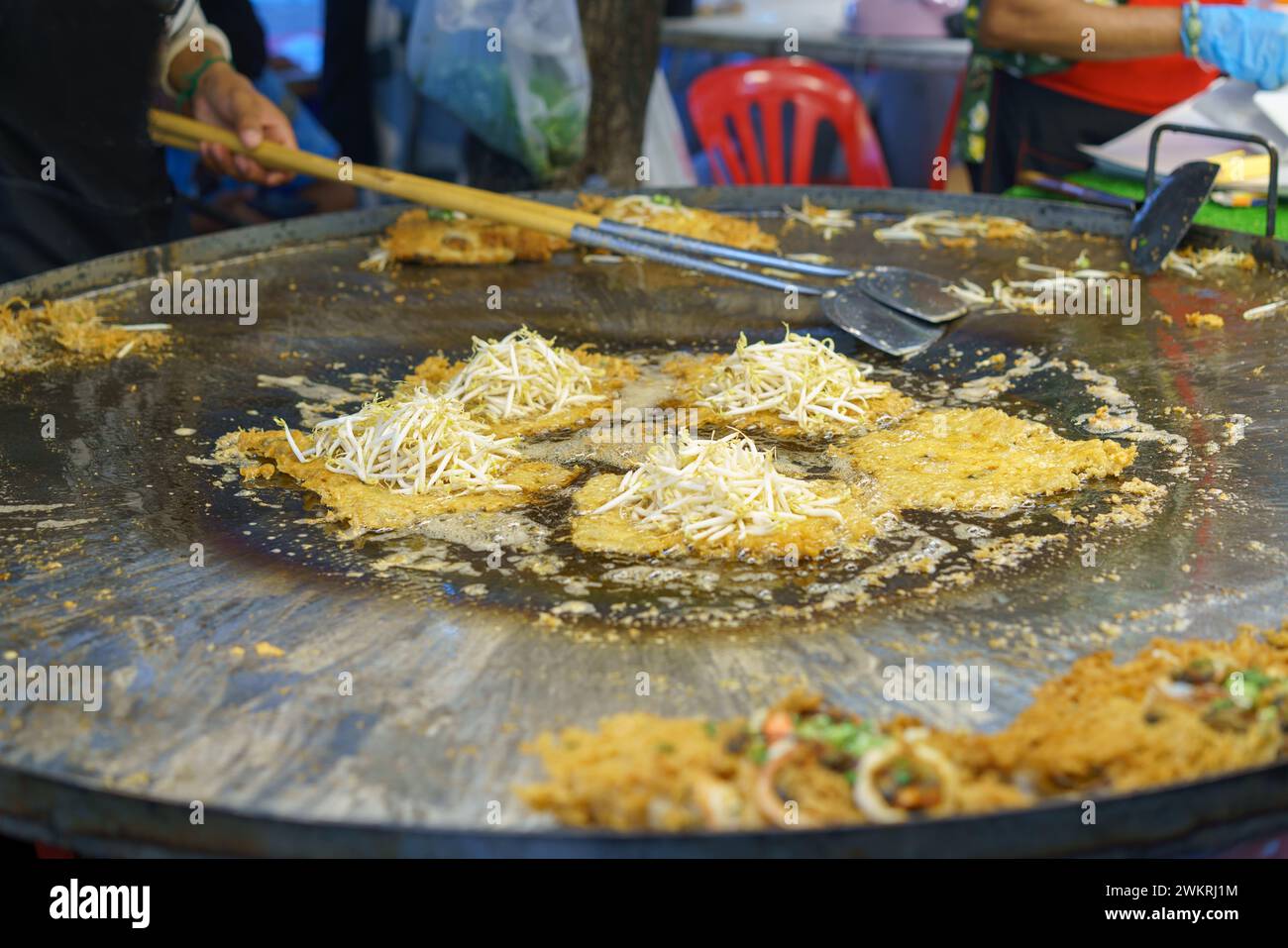 Lo chef thailandese di Street food prepara una tradizionale frittata di ostriche croccanti, con germogli di soia freschi su una grande piastra di ferro Foto Stock