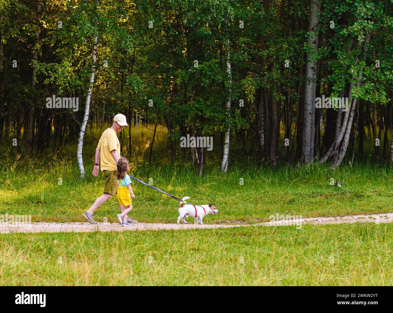 Padre e figlia camminano con un cane da compagnia in una strada di campagna lungo la foresta Foto Stock