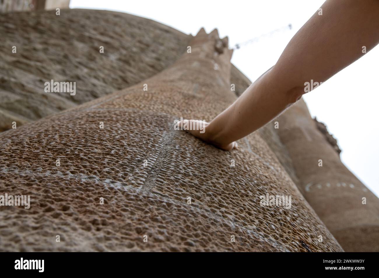 Una mano che tocca una torre della Sagra Familia. La vista va dal basso alla cima della torre. Anche la facciata è in pietra porosa. Foto Stock