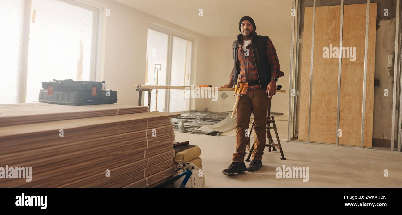 Un imprenditore edile maturo con una cintura portautensili si trova in una cucina, pronto per essere ristrutturato. Questo lavoratore qualificato è specializzato nel miglioramento della casa e nella ristrutturazione Foto Stock