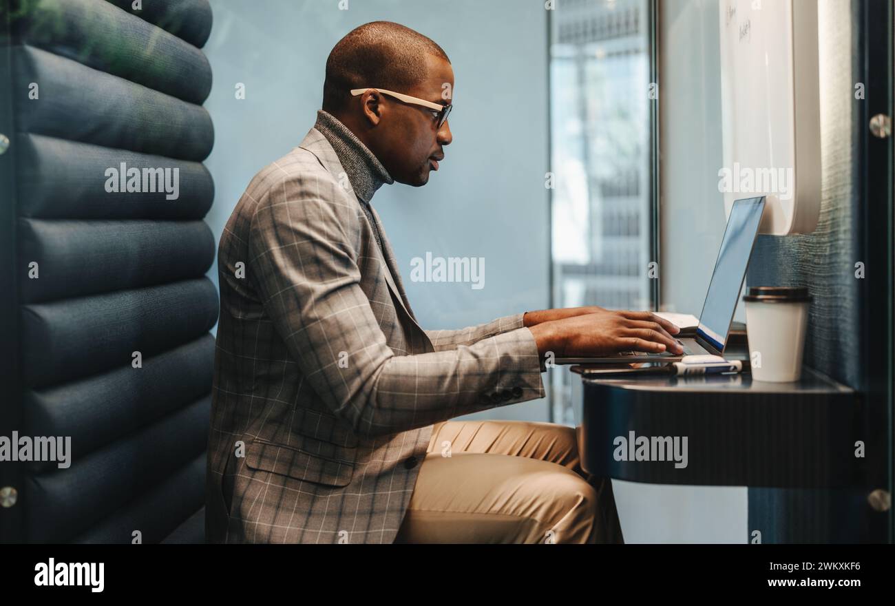 Giovane uomo concentrato con occhiali che lavora su un notebook in un moderno ufficio di coworking. Uomo d'affari che digita diligentemente all'interno del suo stand, creando una professio Foto Stock