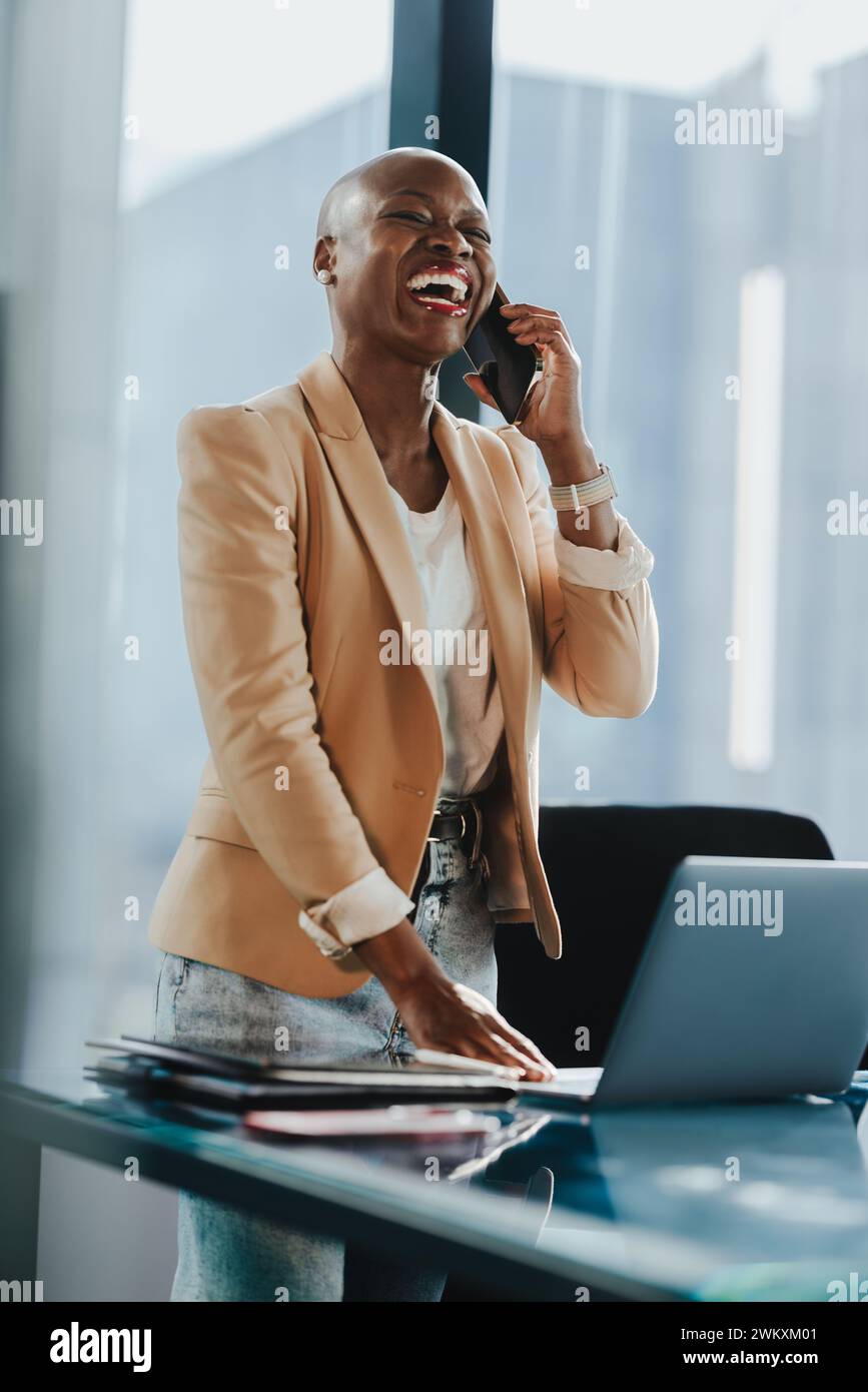 Donna d'affari africana in un ufficio aziendale utilizza il suo smartphone per parlare e discutere di lavoro. Sta accanto alla sua scrivania con un notebook, mostrando un professionista Foto Stock