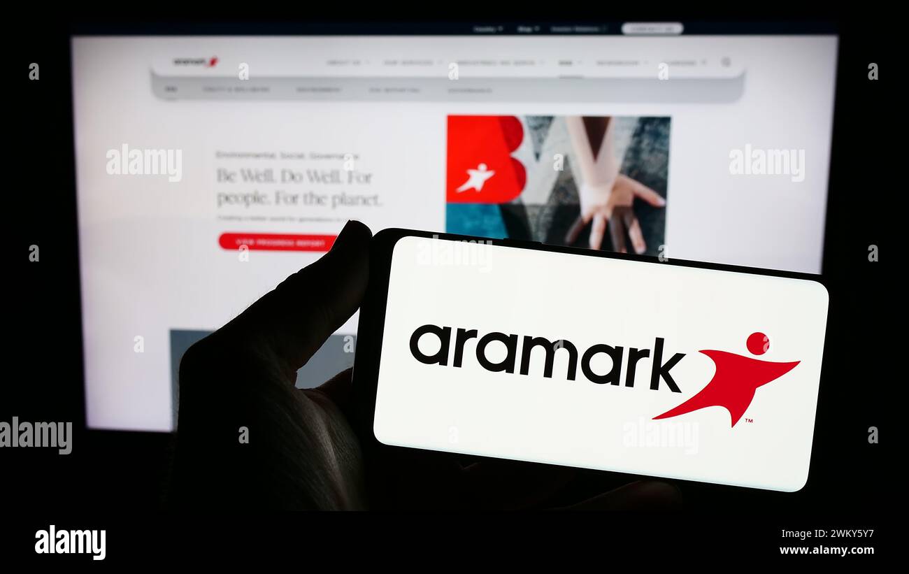 Persona che possiede uno smartphone con il logo della società statunitense di servizi alimentari e gestione di strutture Aramark davanti al sito web. Mettere a fuoco il display del telefono. Foto Stock