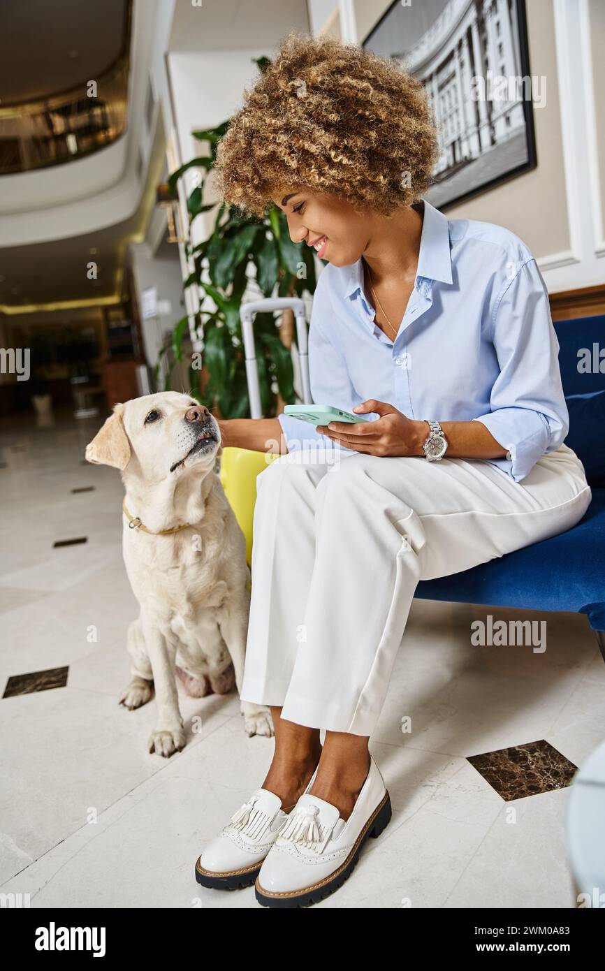 turista con un cane coccolante al telefono in un hotel in cui sono ammessi gli animali domestici, labrador e una donna afroamericana felice Foto Stock