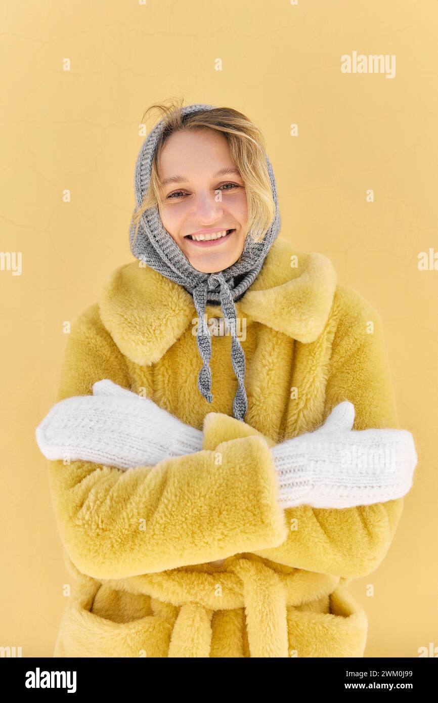 Giovane donna felice che indossa un cappotto di pelliccia e guantoni che sentono freddo davanti al muro giallo Foto Stock
