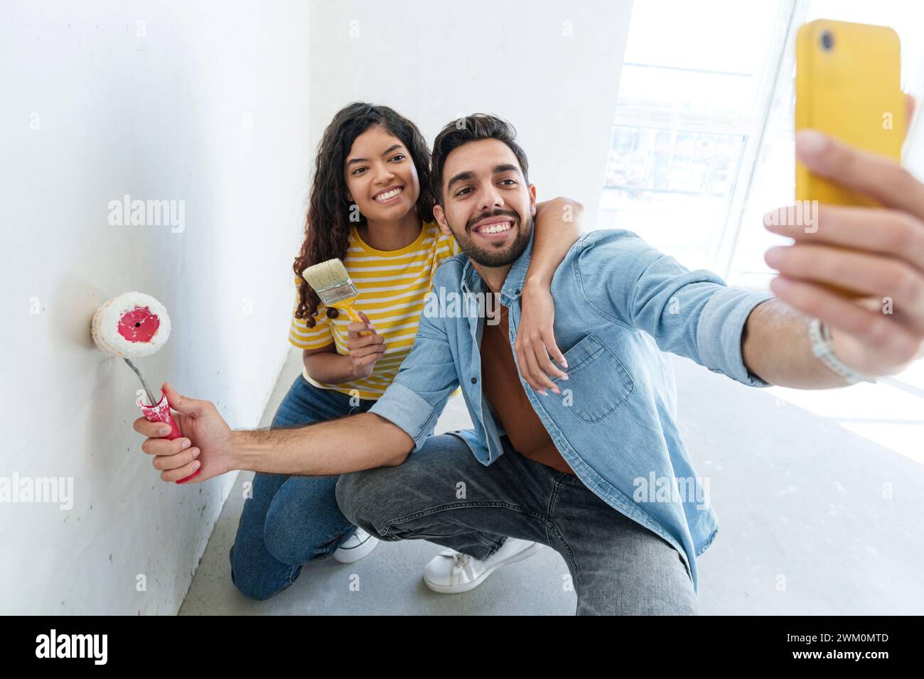 Coppia giocosa con rullo per pittura e pennello che scattano selfie tramite smartphone nella nuova casa Foto Stock