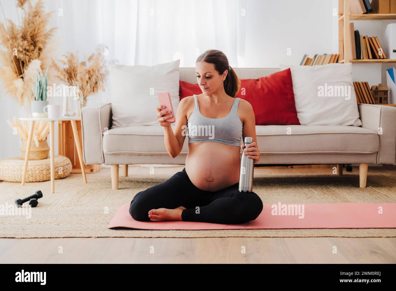 Donna incinta con bottiglia d'acqua che usa il telefono cellulare in soggiorno a casa Foto Stock