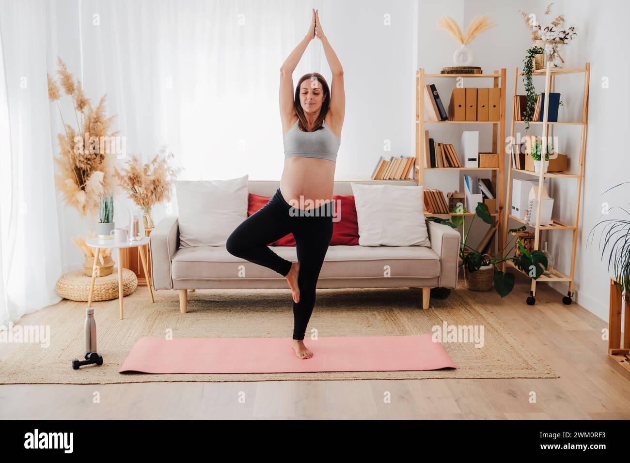 Donna incinta che pratica yoga in posa sugli alberi sul tappetino in soggiorno Foto Stock