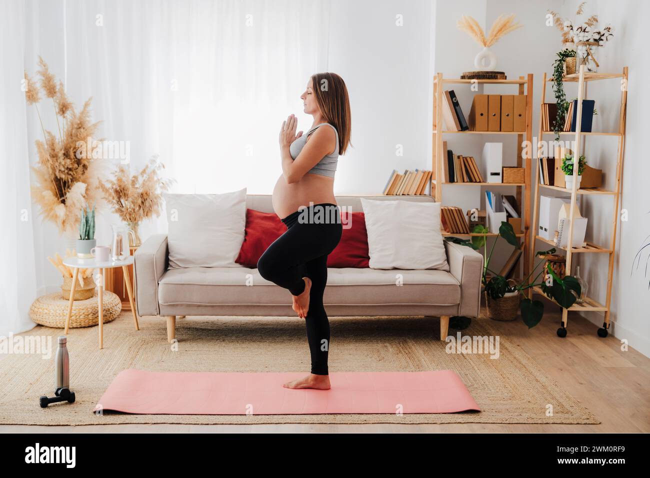 Donna incinta che pratica yoga in posa sugli alberi sul tappetino nel soggiorno di casa Foto Stock