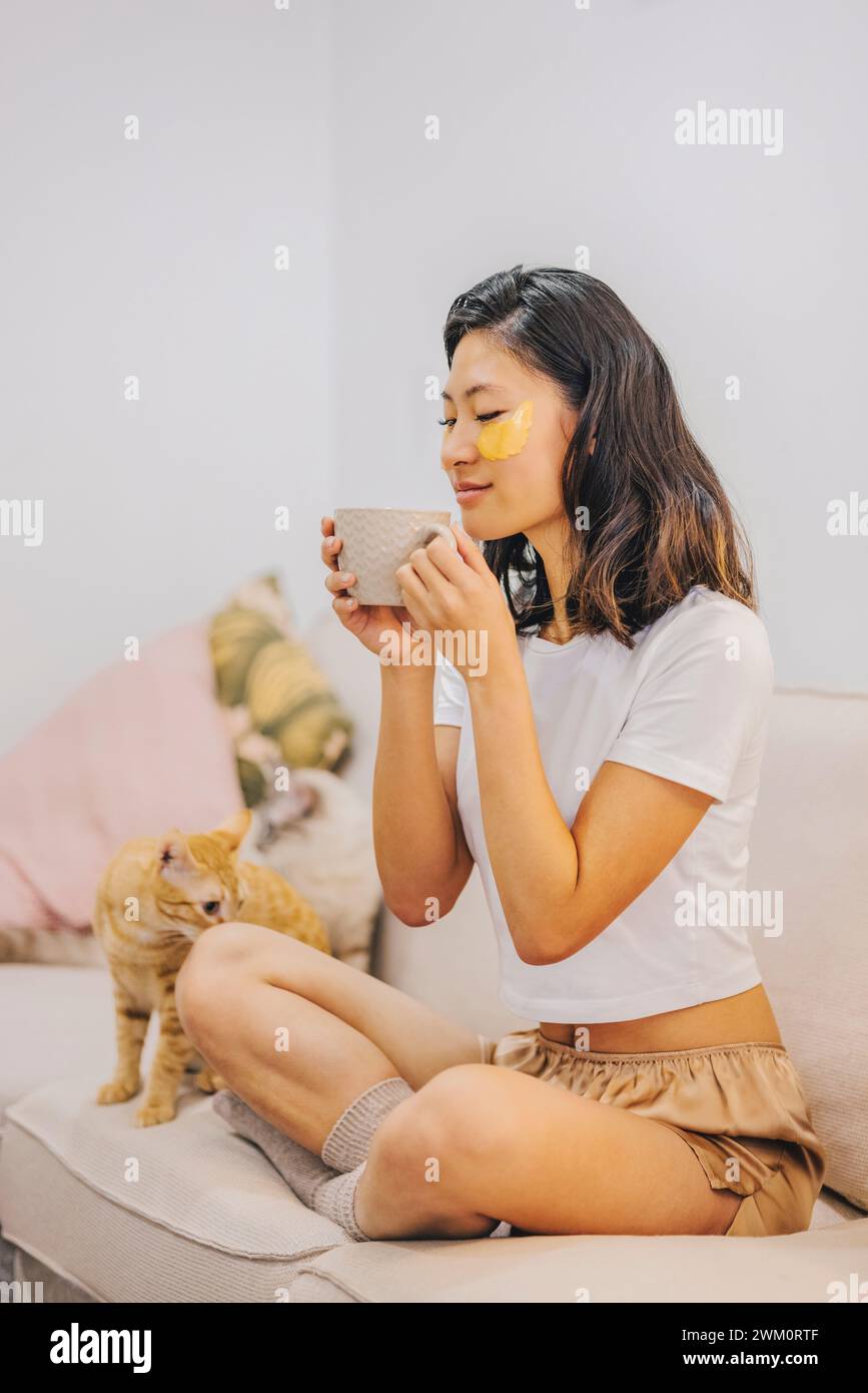 Donna con toppe per la cura della pelle sotto gli occhi che prende il caffè vicino al gatto a casa Foto Stock
