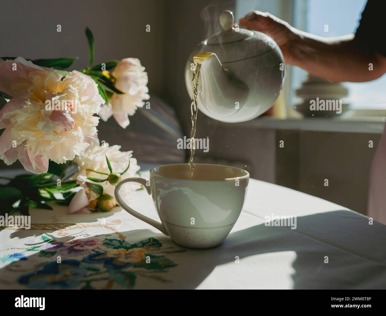 Donna che versa il tè in tazza vicino ai fiori sul tavolo a casa Foto Stock