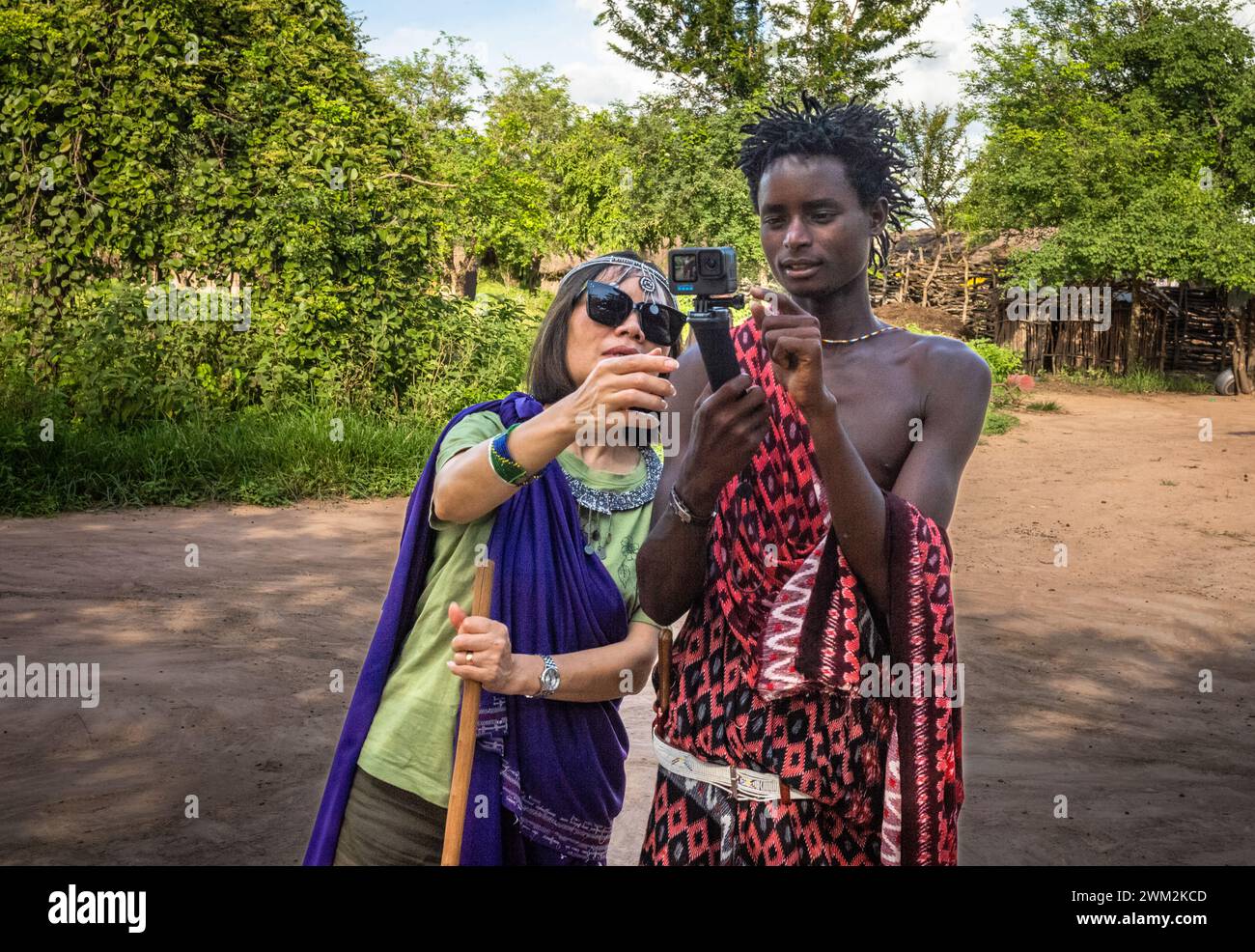 Una donna turistica asiatica mostra a un giovane guerriero Maasai come utilizzare una videocamera GoPro a Mikumi, Tanzania Foto Stock