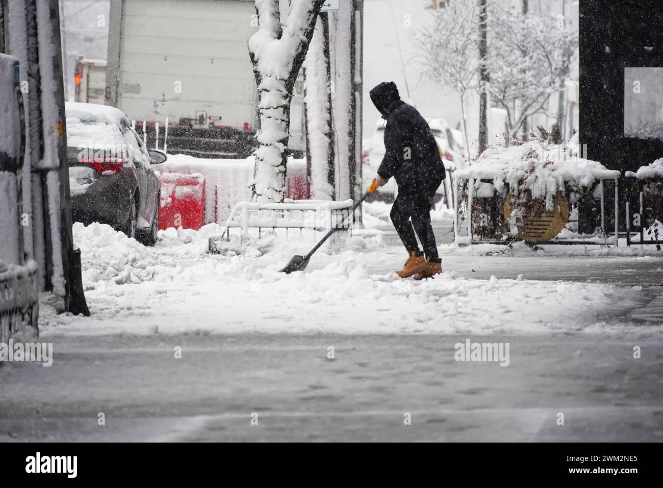 Il proprietario o il dipendente di una piccola azienda all'aperto spalanca il ghiaccio e si bagna sulla strada del centro città con la neve che cade Foto Stock