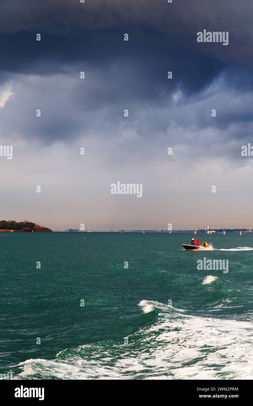 Piccola barca che naviga oltre la Grazia o l'isola di Santa Maria della Grazia nella laguna veneziana in condizioni di tempesta con cielo nuvoloso blu scuro sullo sfondo. Foto Stock