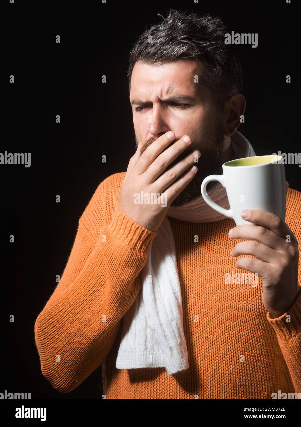 L'uomo tossisce. Maschio malato con raffreddore o bronchite tosse. Bell'uomo barbuto in maglione arancione avvolto in sciarpa con tazza di tè medicinale. Uomo malvagio Foto Stock