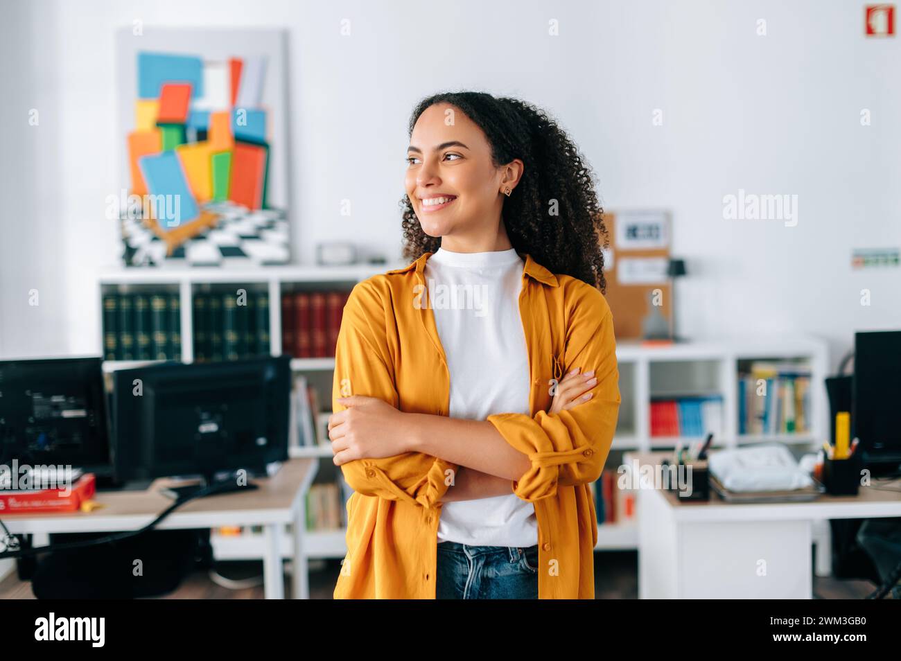 Positivo sicuro di sé bella donna d'affari ispanica dai capelli ricci con camicia arancione, business coach, manager, in piedi in un ufficio moderno con le braccia incrociate, guardando lontano e sorridendo Foto Stock