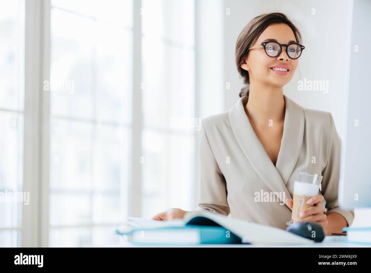 Donna d'affari sorridente con un bicchiere d'acqua in un ufficio luminoso, che promuove il benessere sul lavoro. Foto Stock