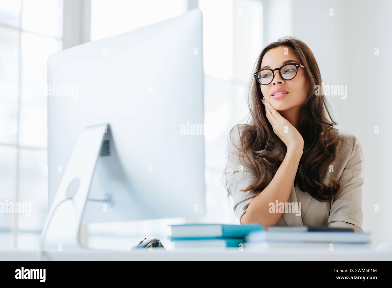 Giovane donna sognante con occhiali persi nel pensiero, che guarda in lontananza in un ufficio luminoso. Foto Stock