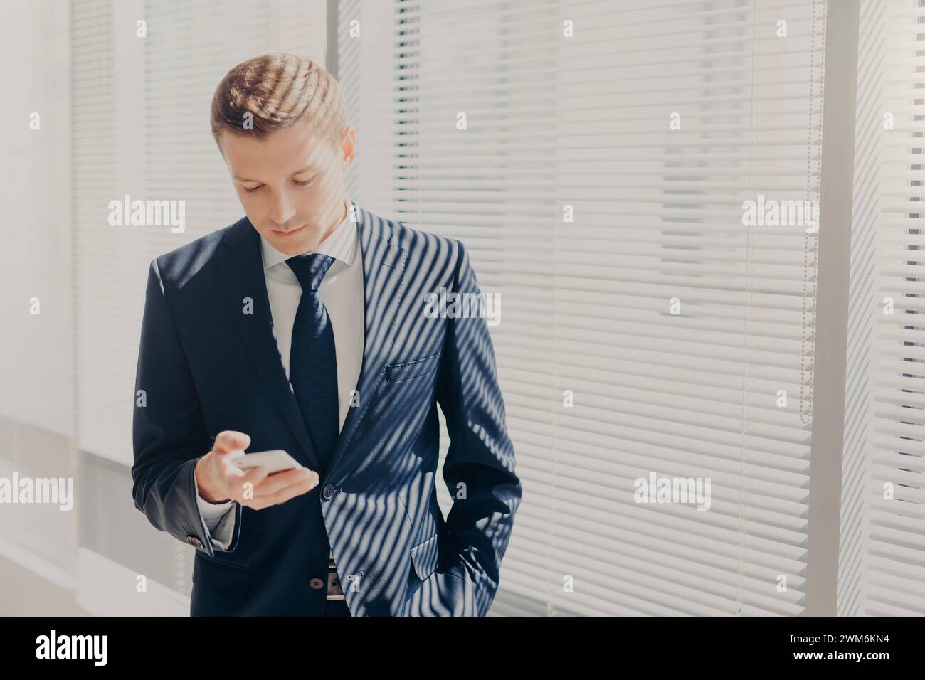 L'elegante uomo d'affari esamina lo smartphone con intenti, stando accanto alle finestre illuminate dal sole. Foto Stock