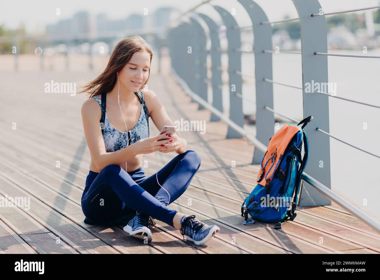 Giovane donna serena seduta su un ponte soleggiato, immersa nel suo smartphone. Foto Stock