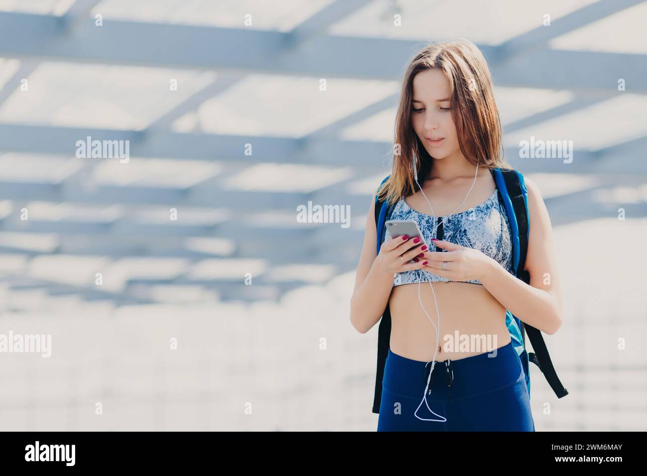 Indossate una giovane donna con uno zaino, controllando il telefono con le cuffie collegate. Foto Stock