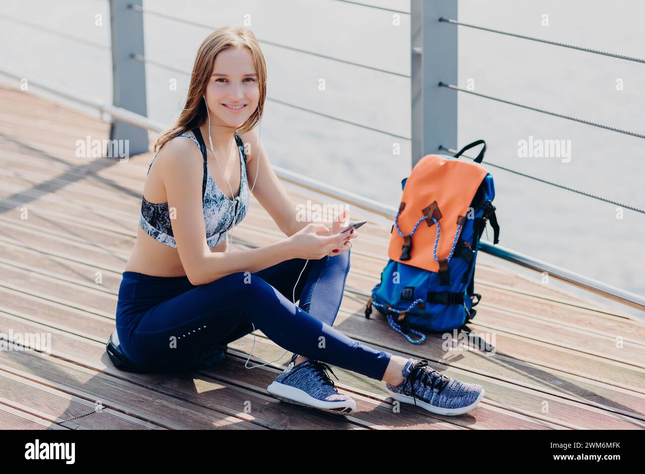 Donna sorridente con gli outfit per il fitness che si ferma per una pausa con lo smartphone sulla passeggiata. Foto Stock