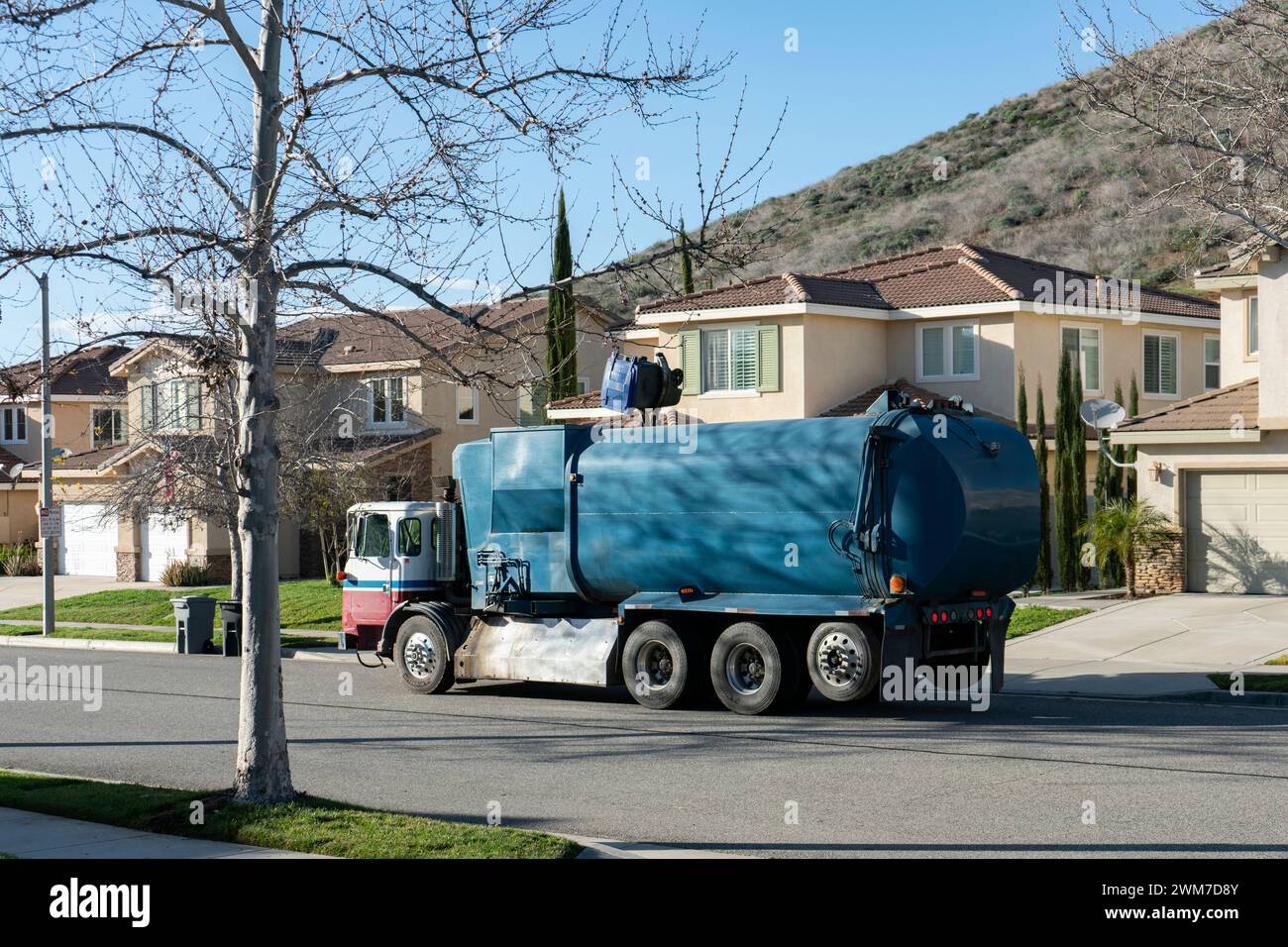 Un camion per la raccolta rifiuti blu raccoglie i rifiuti provenienti dai grandi contenitori per la raccolta rifiuti sul lato di una strada residenziale Foto Stock