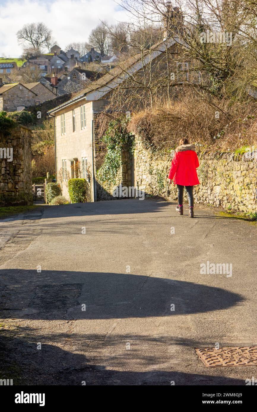 Donna con un cappotto rosso che cammina attraverso il villaggio di Stoney Middleton nel Peak District, passando per i cottage di campagna Foto Stock