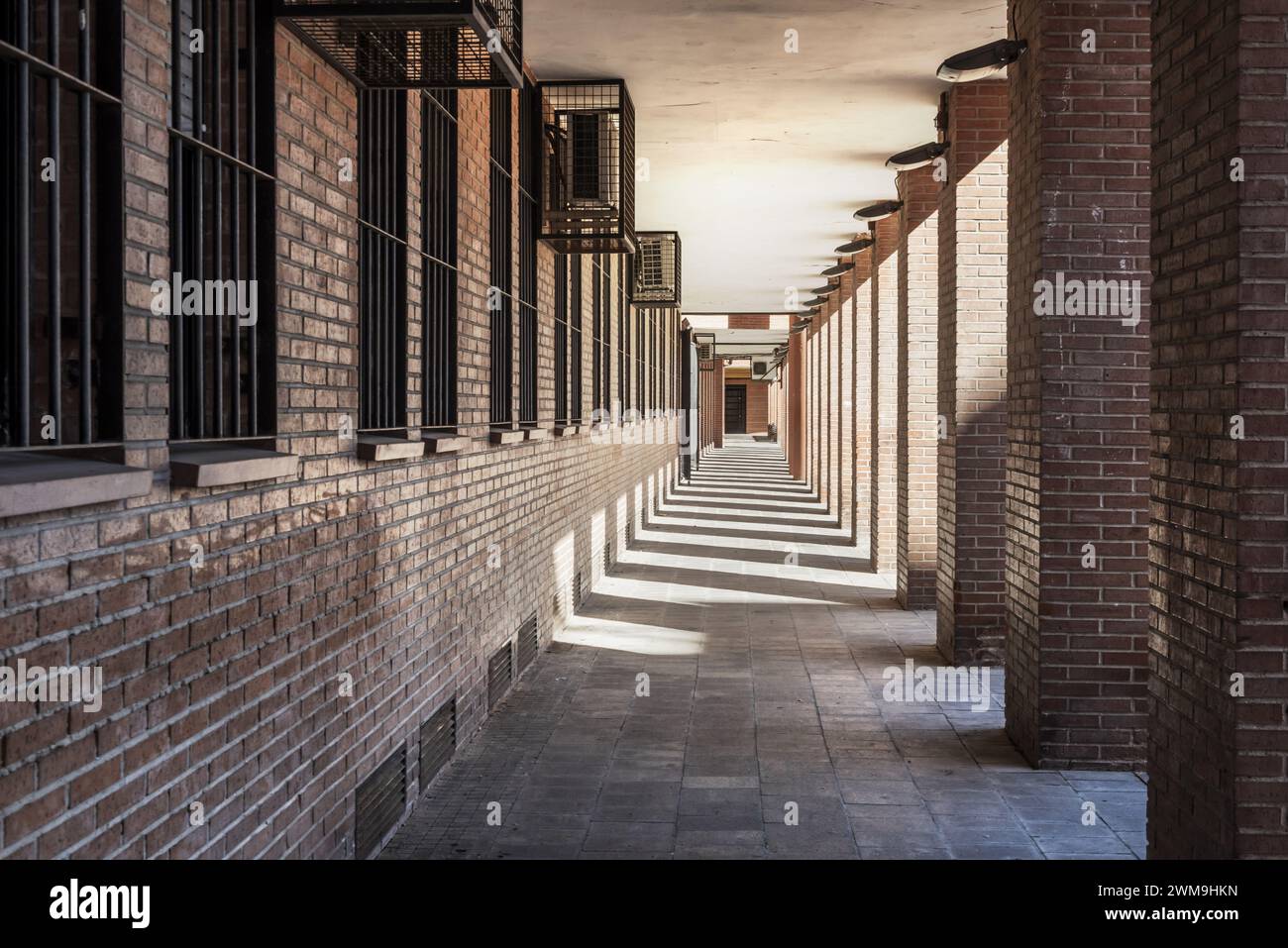 Passaggio al piano terra di un edificio in mattoni marroni con ombre dei pilastri, punto di svanimento e condizionatori d'aria in gabbie metalliche Foto Stock