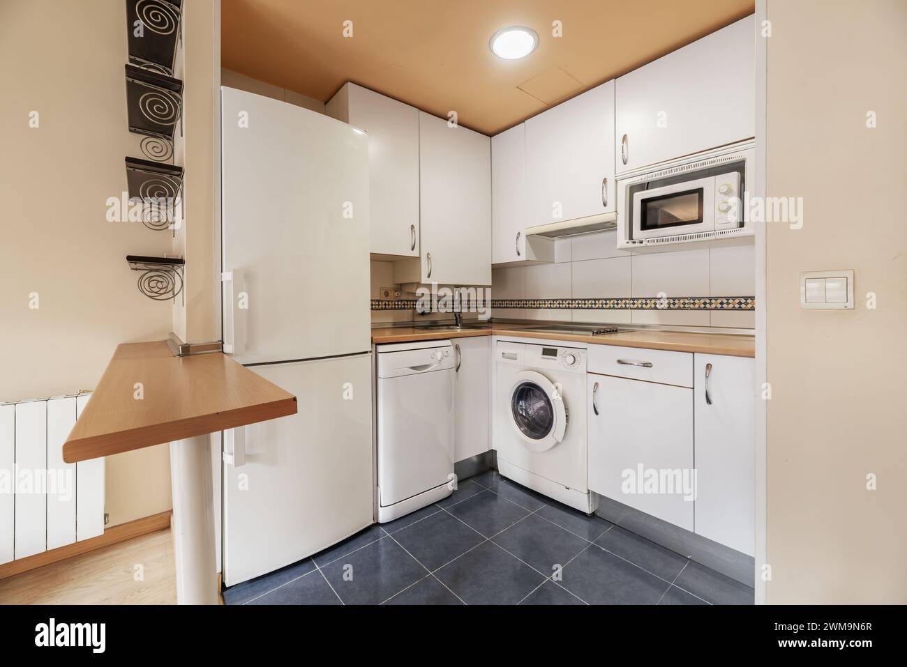 Cucina attrezzata e arredata con mobili bianchi, ripiano marrone, pavimenti in gres blu e un piccolo tavolo isolano con gamba in metallo bianco Foto Stock