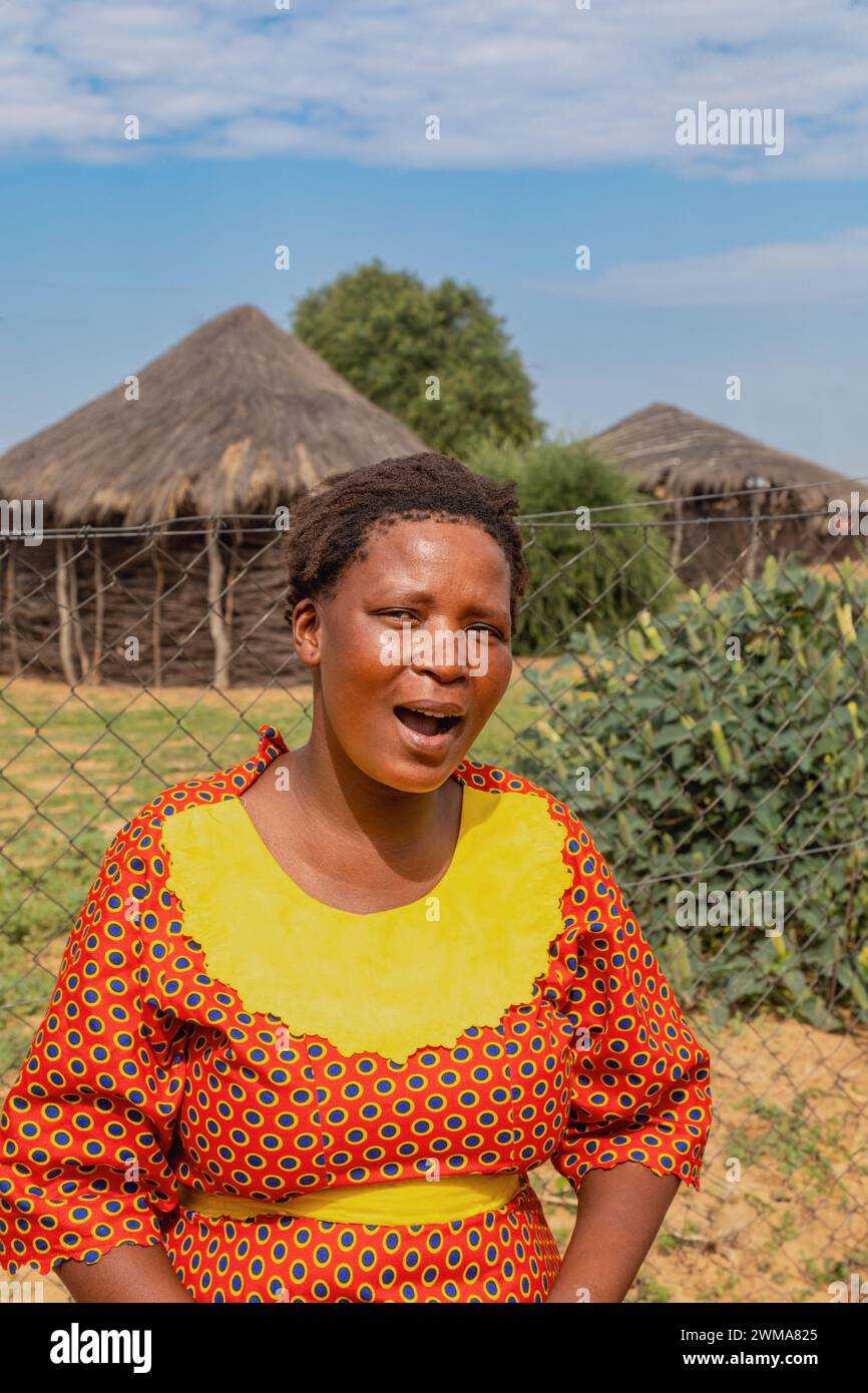 donna africana del villaggio in piedi nel cortile, nella capanna sullo sfondo con tetto di paglia e cielo blu, sud africa Foto Stock