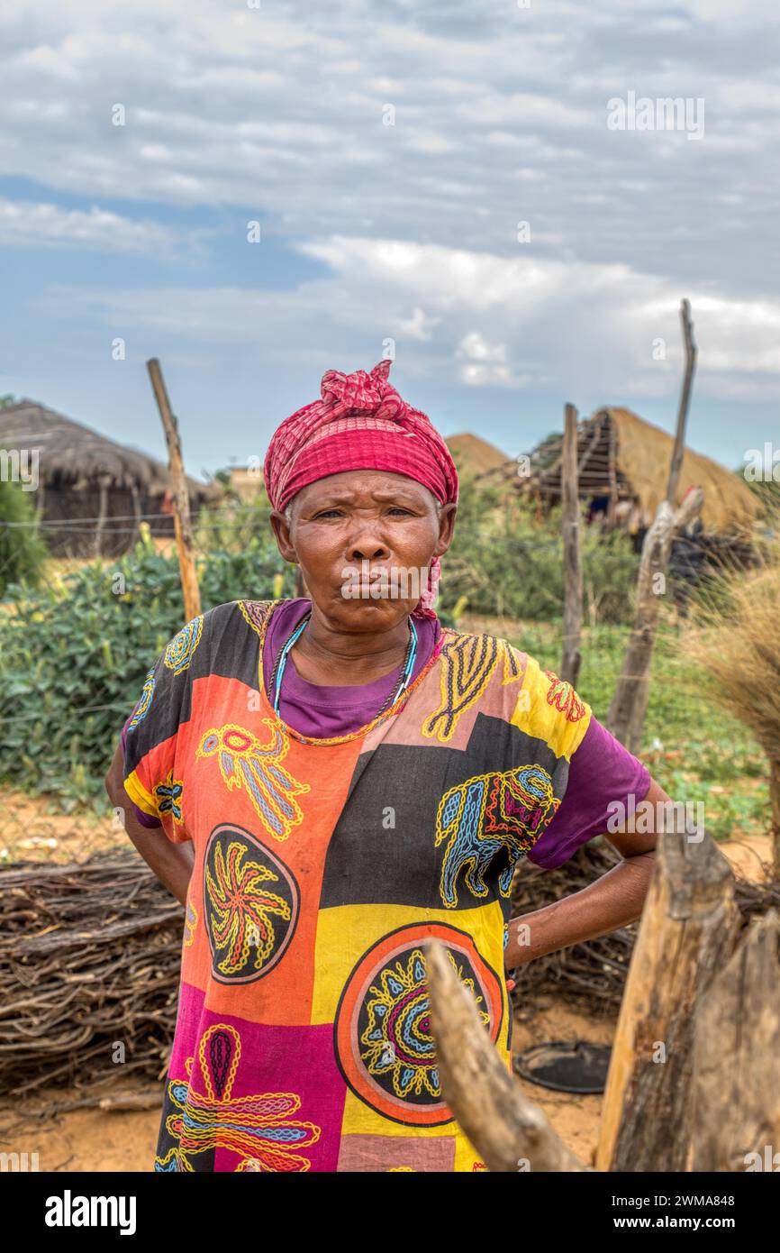 vecchia donna africana del villaggio nel cortile, con collana tradizionale, in baita sullo sfondo con tetto di paglia e cielo blu, sud africa Foto Stock