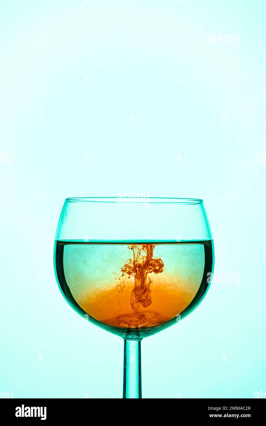 Il colore arancione si mescola con il liquido turchese nel bicchiere da vino su uno sfondo blu uniforme Foto Stock