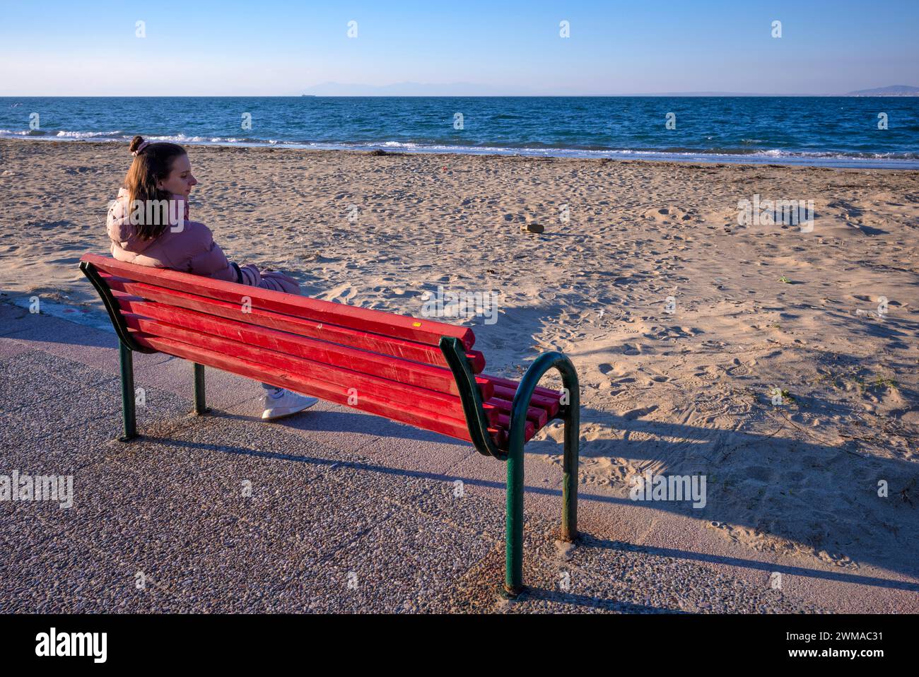 Giovane donna seduta su panchina, panchina, guardando il mare, cappotto, sola, sola, spiaggia, Peraia, anche Perea, luce serale, Salonicco, Macedonia Foto Stock